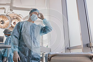 Tired professional surgeon in mask standing in operating room after hard surgery