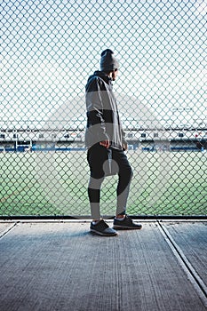 Tired runner athlete holding and looking through rabitz fence on football field. Young sportsman resting after sport excursion run