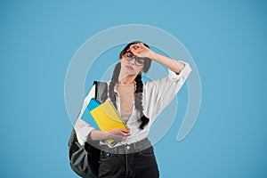 Tired sad teenager european girl with pigtails in glasses with backpack hold books, wipes forehead with hand