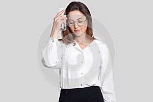 Tired sad woman keeps glass of water near head, feels thirsty and overworked, dressed in stylish formal clothes, isolated over whi