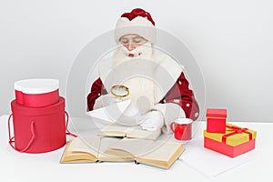 Tired Santa Claus sits at the table and examines letters with a magnifying glass. There are Christmas gifts and books on the table