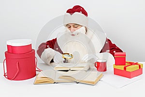 A tired Santa Claus sits at a table and studies books using a magnifying glass. There are Christmas gifts on the table