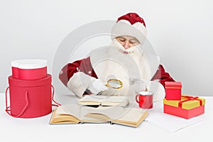 A tired Santa Claus sits at a table and studies books using a magnifying glass. There are Christmas gifts on the table