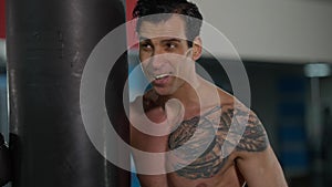 Tired satisfied perspiring sportsman with tattooed chest standing at punching bag looking at camera gesturing. Portrait