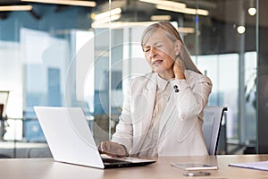 Tired senior gray-haired businesswoman works in office at laptop, massages neck with hand, feels tension and pain