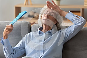 Tired senior woman waving fan feel hot sit on sofa