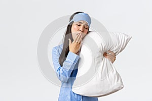 Tired silly asian girl in blue pyjama and sleeping mask, lying in bed and hugging pillow, yawning exhausted, going sleep