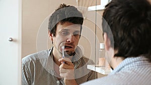 Tired sleepy man with a hangover who has just woken up brush his teeth, looks at his reflection in the mirror.