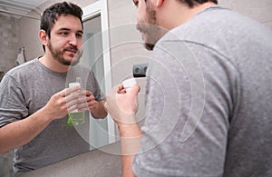 Tired sleepy man rinsing mouth with green mouthwash in bathroom