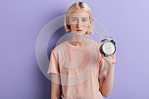 Tired sleepy serious woman holding alarm clock isolated over violet background