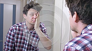 Tired sleepy woken man with a hangover in the bathroom, looks at mirror and puts himself in order.