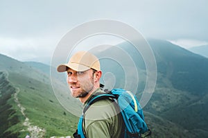 Tired smiling backpacker man in baseball cap walking by the foggy cloudy weather mountain range path with backpack. Active sports