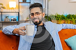 Tired smiling Indian man drinking cup of warm coffee or herbal tea in the morning relaxing enjoying