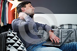 Tired strained man suffering from neck pain while working at computer. Male touchingly massages neck, puts on neck collar, sitting