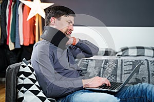 Tired strained man suffering from neck pain while working at computer. Male touchingly massages neck, puts on neck collar, sitting