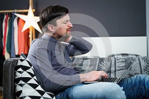 Tired strained man suffering from neck pain while working at computer. Male touchingly massages neck, puts on neck collar, sitting