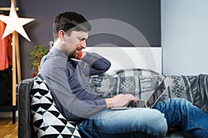 Tired strained man suffering from neck pain while working at computer. Male touchingly massages neck, puts on neck collar, sitting