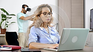 Tired and streesed woman works on the computer while her boyfriend wears virtual reality headset in the background