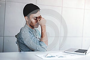 Tired and Stress Young Businessman Sitting on Desk in Office with Computer Laptop. Man Massaging a Nose and Closed Eyes, Keeping