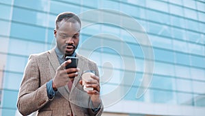Tired and stressed African American businessman reads something in his smartphone standing outside. Man sms texting