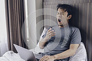 Tired stressed young Asian man feeling sleepy and tired while using laptop on the bed in bedroom. Hard work concept