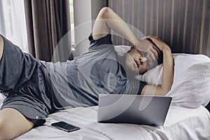 Tired stressed young Asian man feeling sleepy and tired while using laptop on the bed in bedroom. Hard work concept