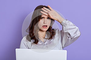 Tired student with headache posing isolated over lilac background and working with portable computer, looks at camera with