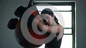 Tired and sweaty woman fighter leaning on the punching bag after intensive trining and looking in the camera.