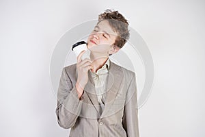 Tired teen boy in grey business suit with paper Cup of coffee in hand on white studio background