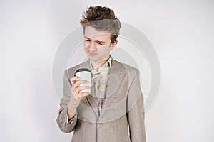 Tired teen boy in grey business suit with paper Cup of coffee in hand on white studio background