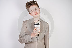 Tired teen boy in grey business suit with paper Cup of coffee in hand on white studio background