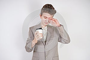 Tired teen boy in grey business suit with paper Cup of coffee in hand on white studio background