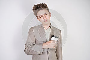 Tired teen boy in grey business suit with paper Cup of coffee in hand on white studio background