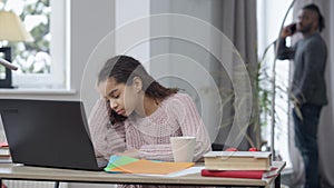 Tired teen student falling asleep listening online lesson on laptop as man walking at background talking on smartphone