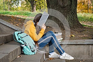 Tired teenage girl reads a book, covers her face with a book sitting on the stairs in the park.