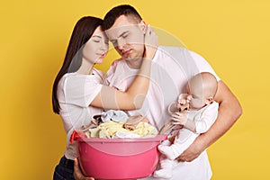 Tired troublesome parents crying newborn baby and basin full of clean clothing, have much domestic work, posing isolated on yellow