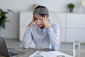 Tired upset unhappy young indian lady suffering from headache at workplace in office interior