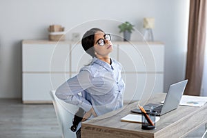 Tired upset young indian business woman suffering from back pain, muscle spasm at workplace with laptop