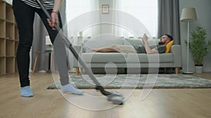 Tired wife vacuuming parquet floor and her husband play on smartphone on sofa