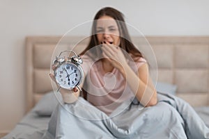 Tired woman in bed with alarm clock, yawning early in the morning