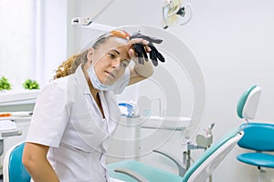 Tired woman dentist, background dental office chair. Female dentist taking off gloves wiping her forehead