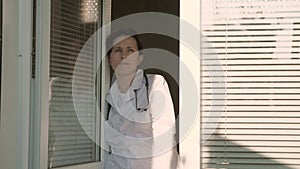 A tired woman doctor in protective suit looks out window, takes off his medical mask, closing his eyes from fatigue