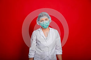 Tired woman doctor with a white coat, mask and medical cap is standing on a red background.