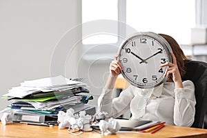 Tired woman hiding face behind clock at table in office. Time management concept