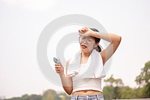 A tired woman holds a small fan and wipes her sweat on her face while walking outdoors on a hot day