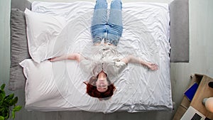 tired woman lies on the bed with her arms outstretched to sides in bedroom, top view