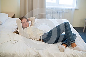 Tired woman is lying in bed with her arm on head and eyes. Young woman with long hair, wears jeans