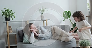 Tired woman lying on couch relaxing cosiness home interior. Cute little boy kid running having fun