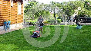 Tired woman mowing green lawn in backyard near house and car moving along area with lawn mower on summer sunny day, slow