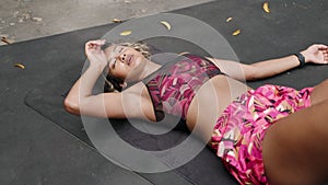 Tired woman rests on mat after intense outdoor workout session. Biethnic female in fitness attire, lying down